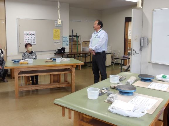 広々とした陶芸教室で、男性講師が白いシャツに黒いパンツ姿で立ち、複数のテーブルに分かれて座っている生徒たちに向けて説明をしている様子の写真