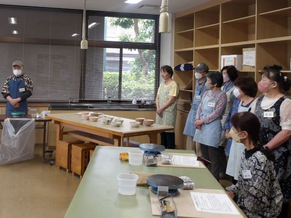 ろくろや道具が並べられた陶芸教室で、複数の生徒がエプロン姿で講師の説明を聞いており、窓からは明るい光が差し込んでいる様子の写真
