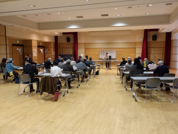 多目的ホールで講師が前に受講者とスピーカーから流れる音楽をきいている