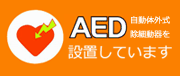 オレンジ色の背景に、左側に白い円の中に雷のような模様が入った赤いハートのイラストがあり、右側には大きな文字で「AED」と、その下に「設置しています」、右側に小さく「自動体外式除細動器を」と書かれている、AED設置を知らせる看板