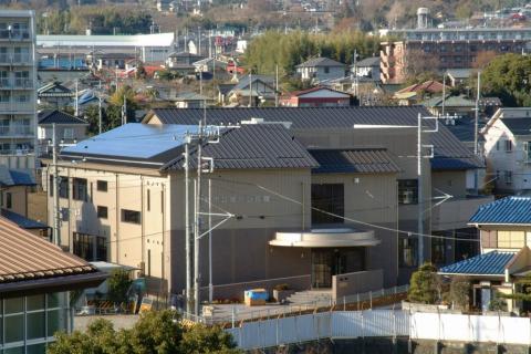 住宅が密集する地域に建つ、茶色とベージュのツートンカラーの現代的な外観の建物で、一部の屋根にはソーラーパネルが設置されており、手前には円形の屋根を持つエントランスがある風景の写真