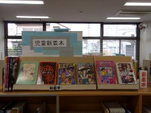 図書館の児童新着本コーナーに、青と白で「児童新着本」と書かれたボードが飾られ、様々なテーマの児童書が平積みと面陳で展示されている様子を捉えた写真