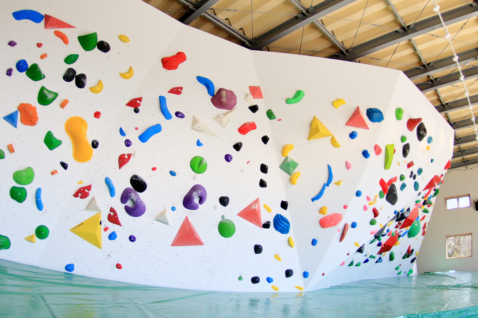 白い室内ウォールいっぱいに様々な色と形をしたカラフルなクライミングホールドが取り付けられており、手前には落下時の安全を確保するための緑色の分厚いマットが敷かれている写真