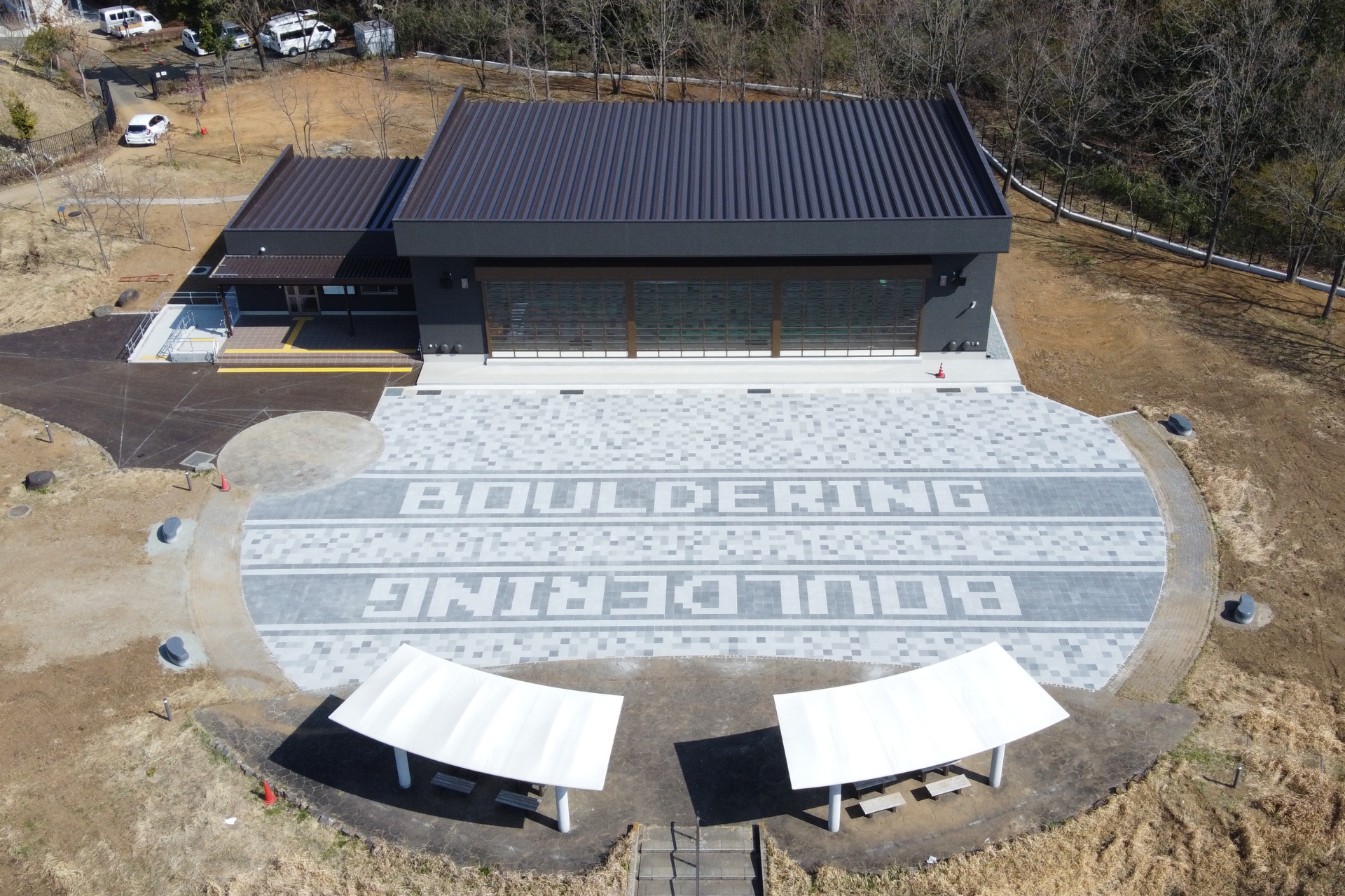 広大な敷地の中央に建つ、黒い外壁と屋根を持つ平屋の建物と、その前の楕円形の広場に「BOULDERING」の文字がタイルでデザインされた屋外施設の俯瞰写真