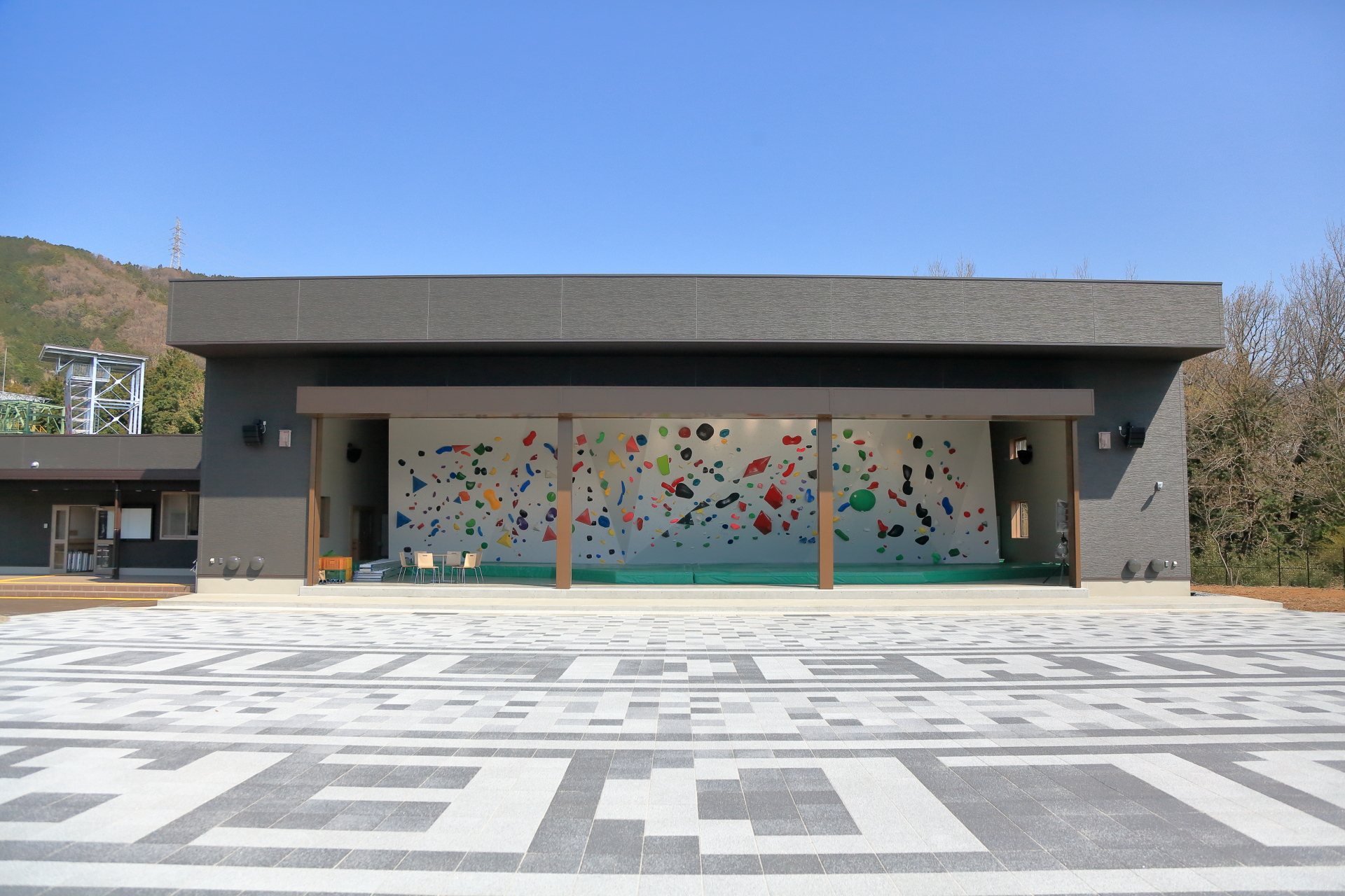 低層でモダンなデザインの建物の前面に、様々な色と形のホールドが配置された大きなクライミングウォールが開口部に設置されており、手前には幾何学模様のタイルが敷き詰められた広場が広がっている写真