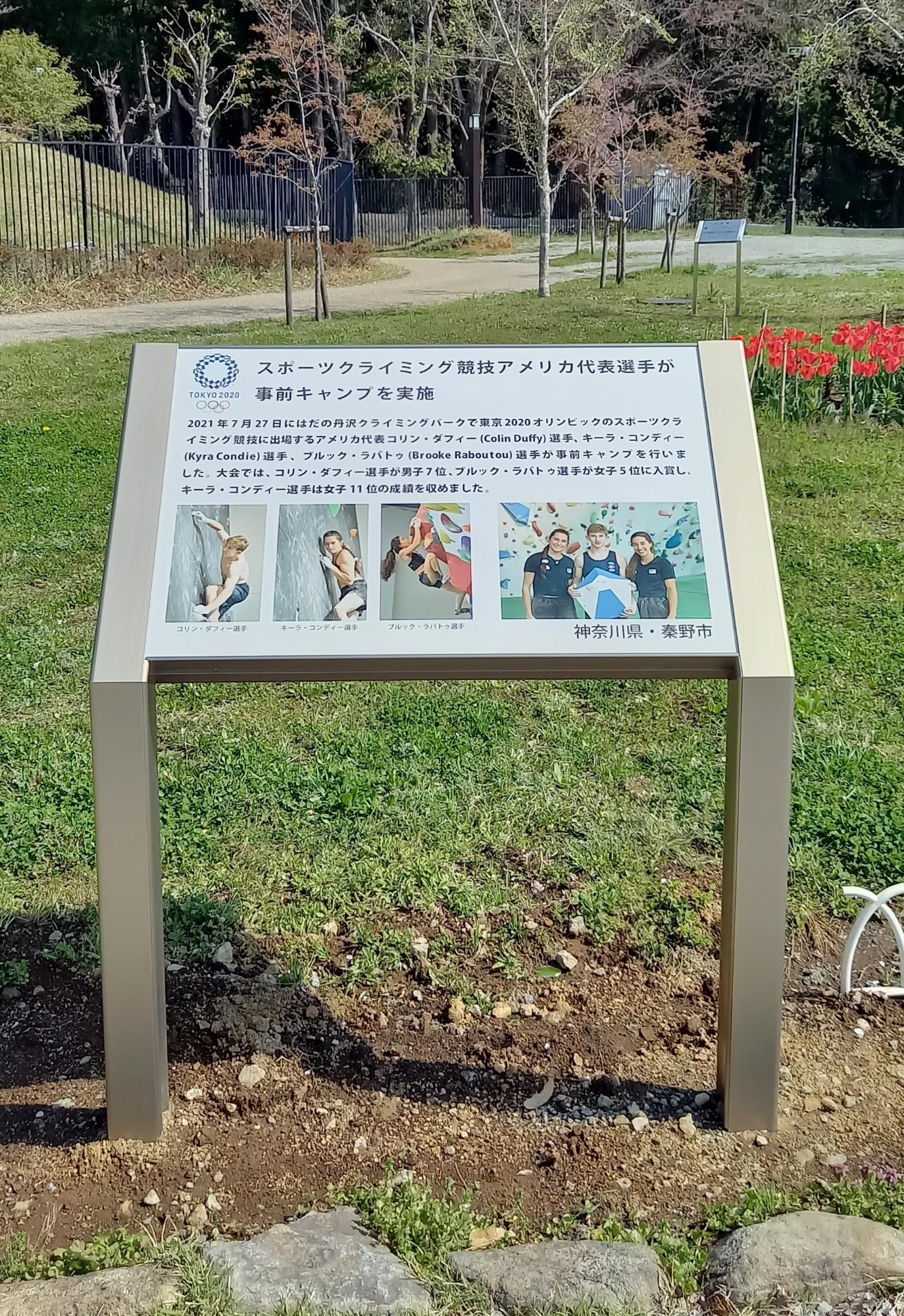 野外の芝生の中に設置された、「スポーツクライミング競技アメリカ代表選手が事前キャンプを実施」と題し、東京2020オリンピック関連の選手と成績を紹介する内容が記載されている説明看板を縦向きに撮影した写真