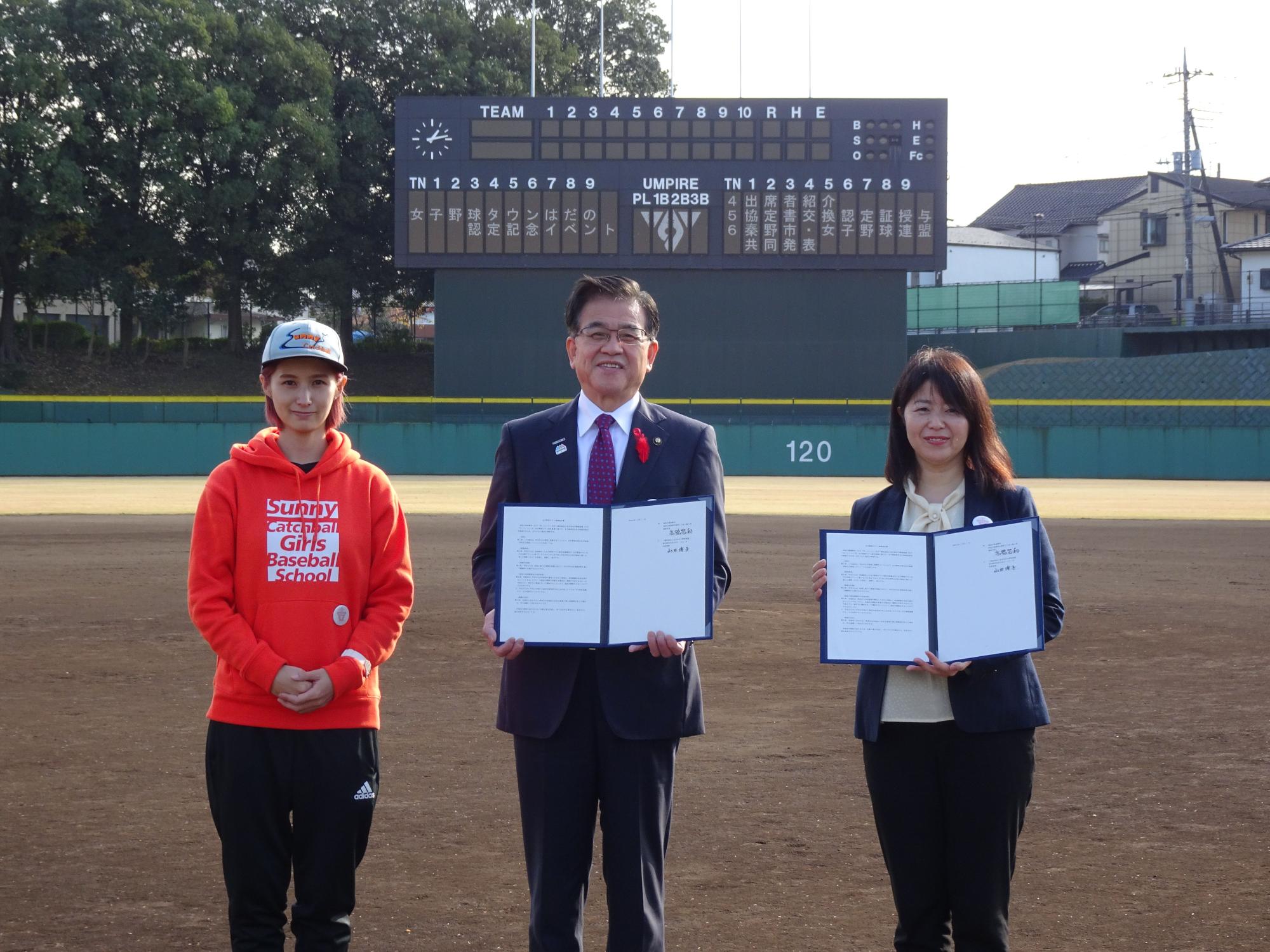 屋外の野球場で、、チーム名やイニング、スコアなどが表示される大きな電光掲示板を背景に、オレンジ色のパーカーとキャップを被った女性の横に立った市長と黒いジャケットの女性が認定証を広げて立って写した記念写真