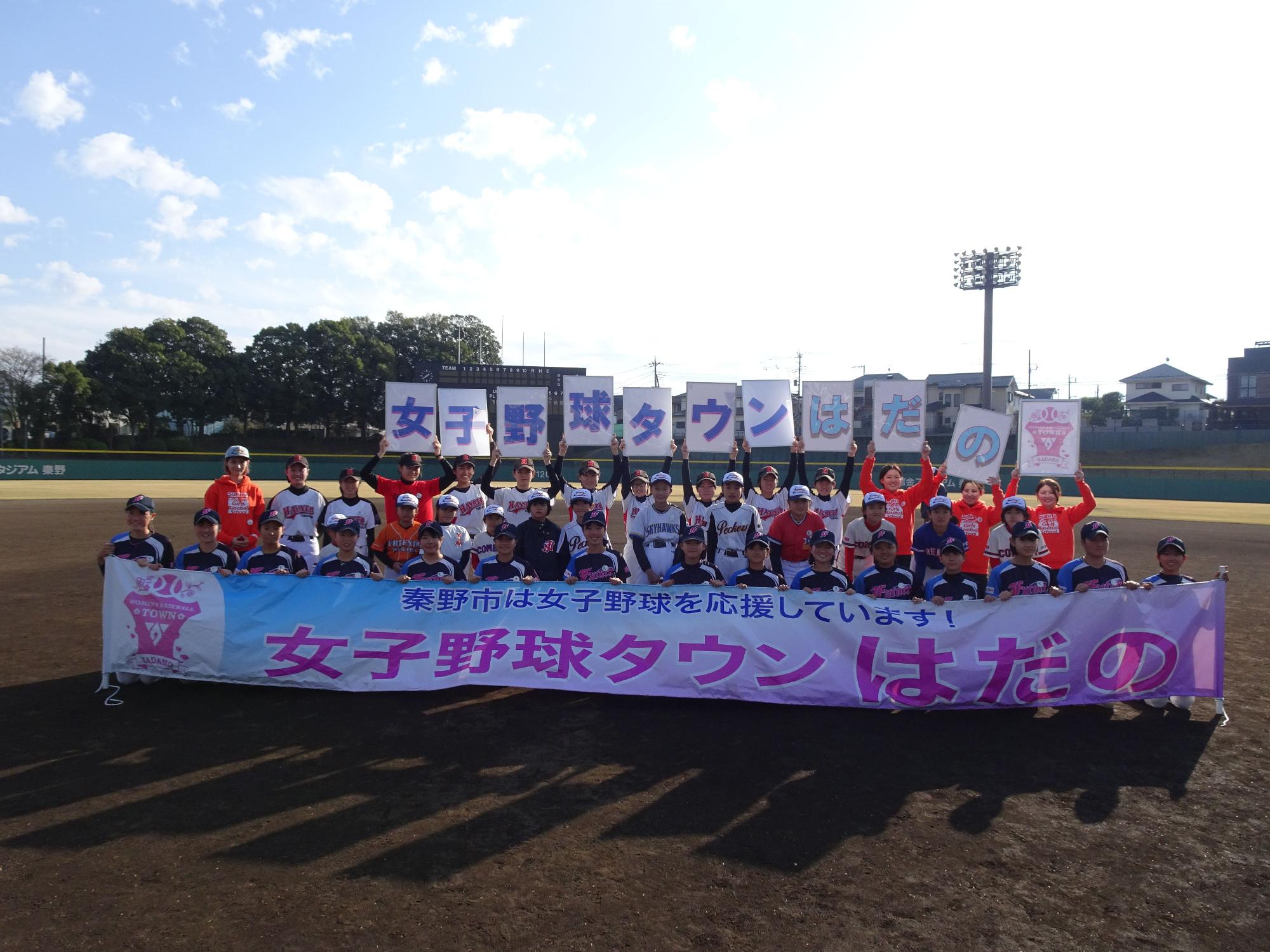 ユニフォームやオレンジ色のパーカーを着た大勢の子ども、大人が、晴天の野球場のグラウンドで、「女子野球タウン はだの」と書かれた横断幕と文字パネルを掲げて写した集合写真