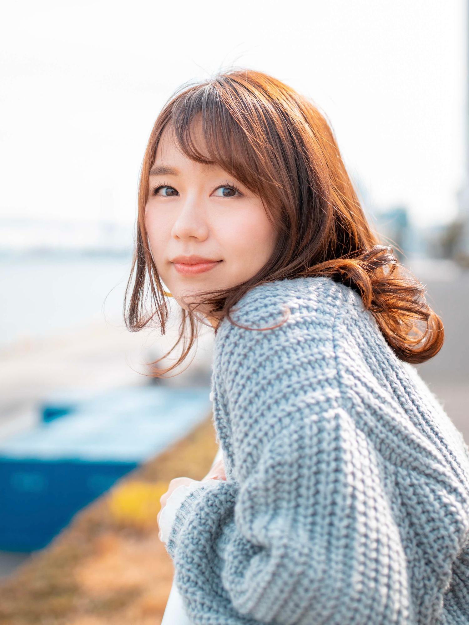 暖色系の光に照らされた茶色の髪の立石純子さんが、ケーブル編みのグレーのセーターを着て、川や海沿いの柵にもたれかかり、横向きからカメラに視線を送っている写真