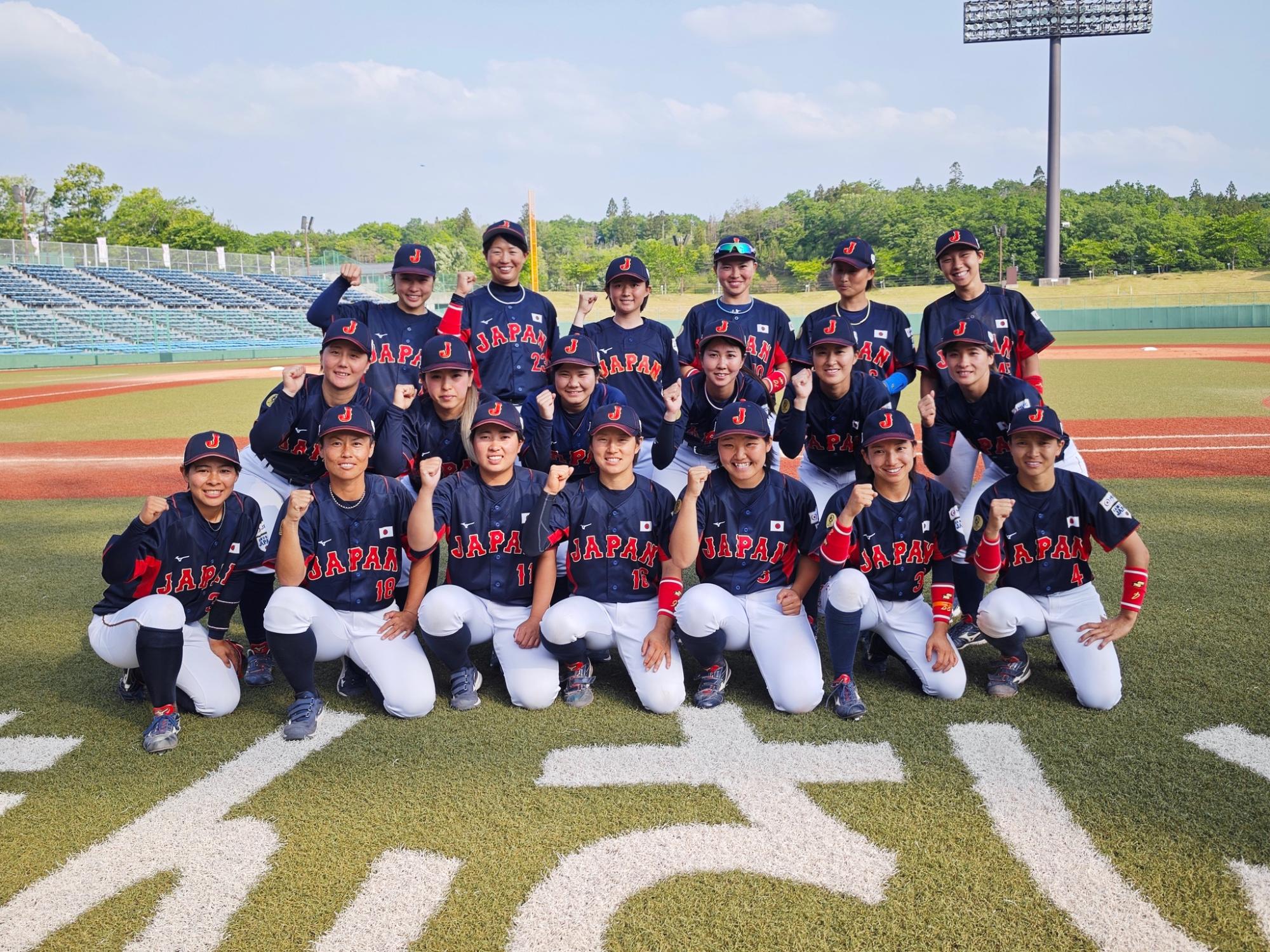 ソフトボール日本代表のユニフォームを着た18人の選手が、晴れた日の野球場の芝生の上で、前列がしゃがみ後列が立って並び、皆が拳を握り笑顔でカメラ目線で勝利を誓う集合写真