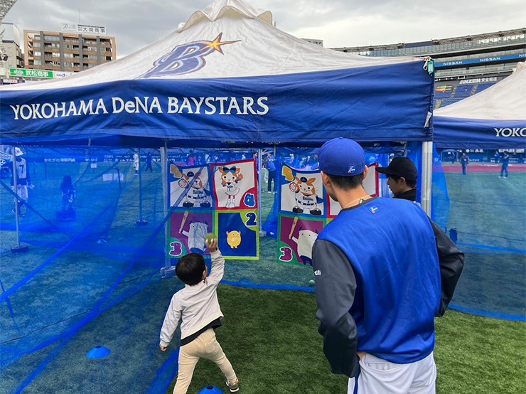 スタジアムの外野スペースにある青いテントの下で、横浜DeNAベイスターズのチーム名のロゴが入ったブルーのテントの下、小さな男の子が野球の投球ゲームに挑戦しており、野球帽をかぶった男性がその様子を見守る、ファンサービスイベントの風景写真