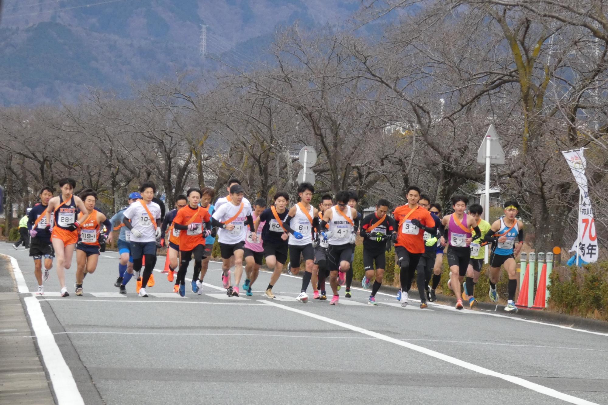 多数のランナーが参加するマラソン大会のスタート直後か序盤の様子を捉えた写真で、選手たちが車道のアスファルトの上をオレンジ色や白のウェアを着て一斉に走り出している写真