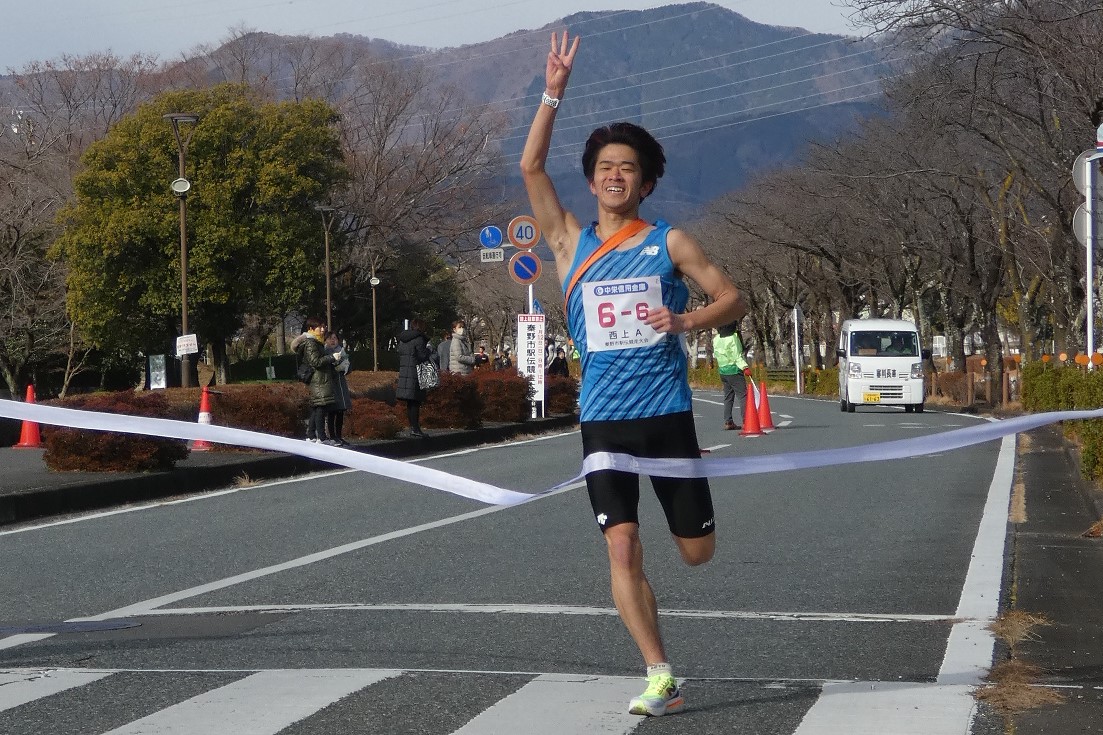 マラソン大会で青いタンクトップと黒いランニングショーツを着用した若い男性ランナーが、ガッツポーズをしながら白いテープのゴールラインを笑顔で通過する瞬間の写真