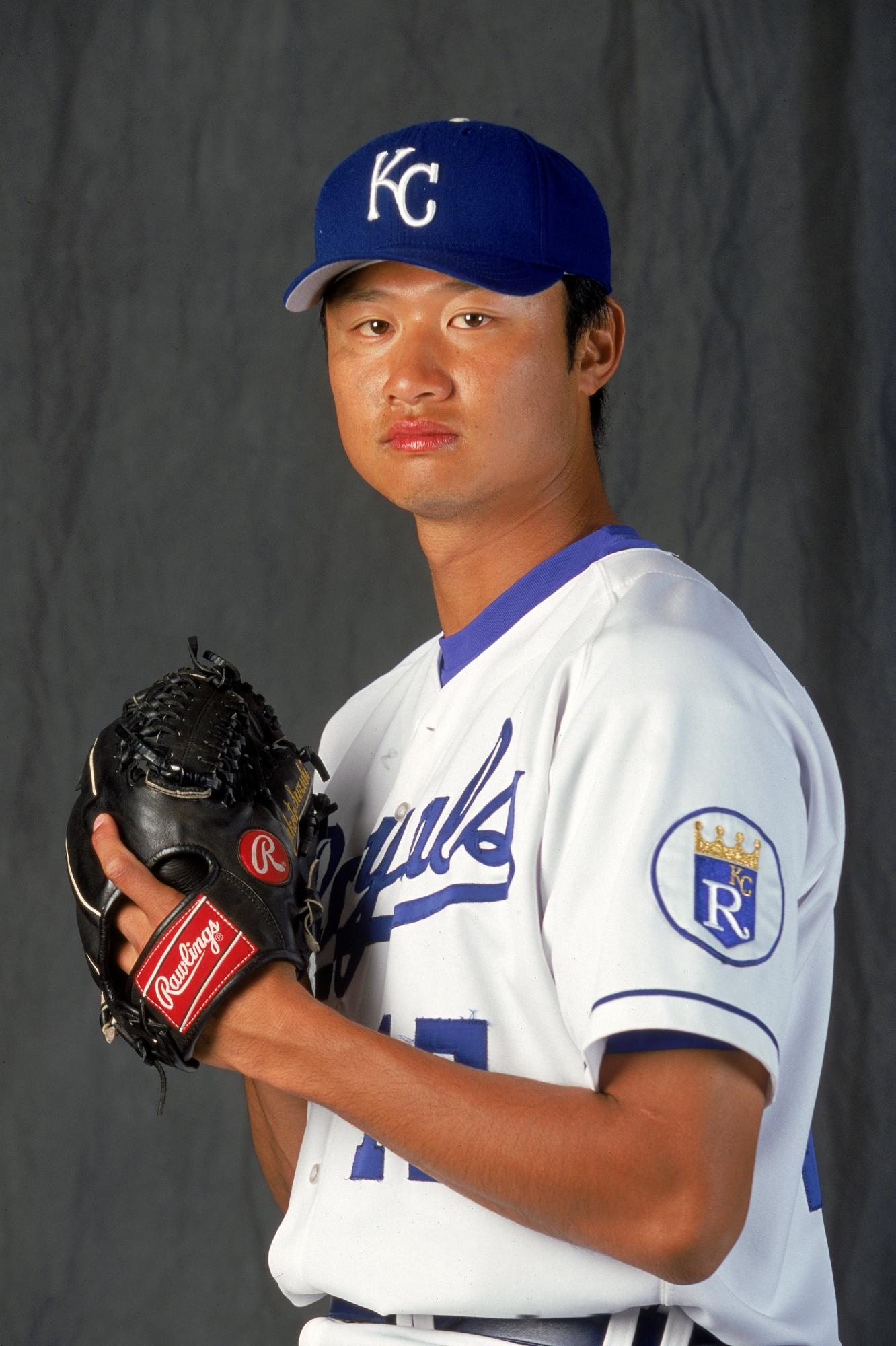 MLBカンザスシティ・ロイヤルズのユニフォームを着たアジア系のマック鈴木氏が、青いキャップと白いユニフォーム姿で、左手に黒いローリングス社のグローブをはめてカメラをまっすぐに見つめている上半身のポートレート写真