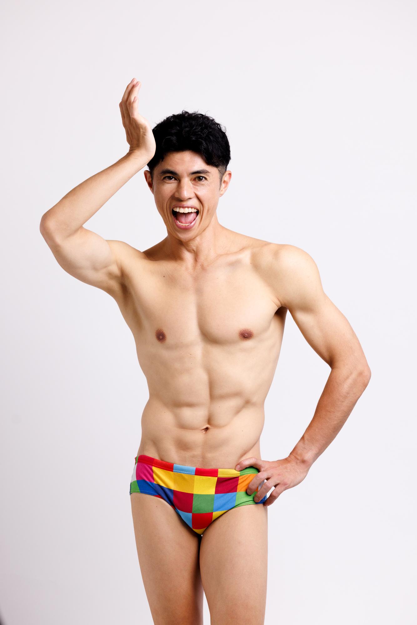 白い背景の前で、カラフルなチェック柄のブリーフ型水着を着用した引き締まった体格の小島よしおさんが、片手を頭の上に掲げてもう一方の手に腰を当て、口を大きく開けて歓声を上げている様子の写真