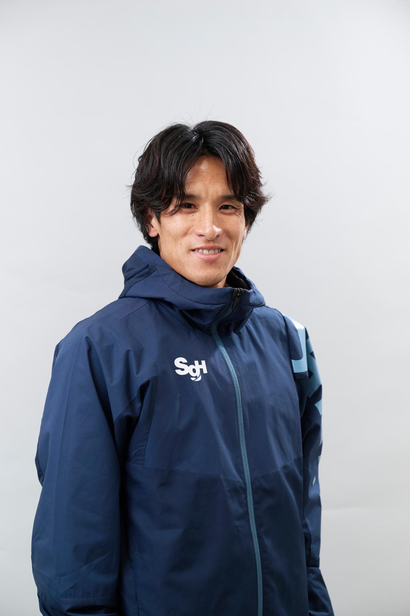 白い背景の前で、黒い髪の村澤明伸さんが濃紺色のフード付きジャケットを着用し、カメラに向かって穏やかに微笑んでいる、上半身を写した写真