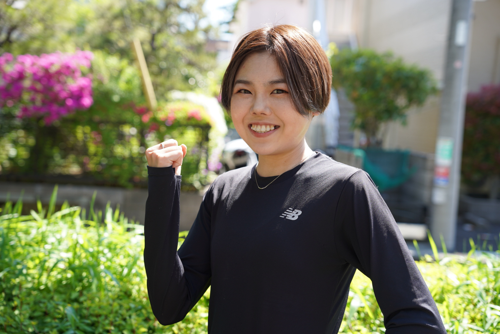 短い茶髪の萩谷楓さんが、黒い長袖のスポーツウェアを着用し、明るい日差しの屋外で、右手の拳を力強く握りしめてカメラに向かって満面の笑みを浮かべている写真