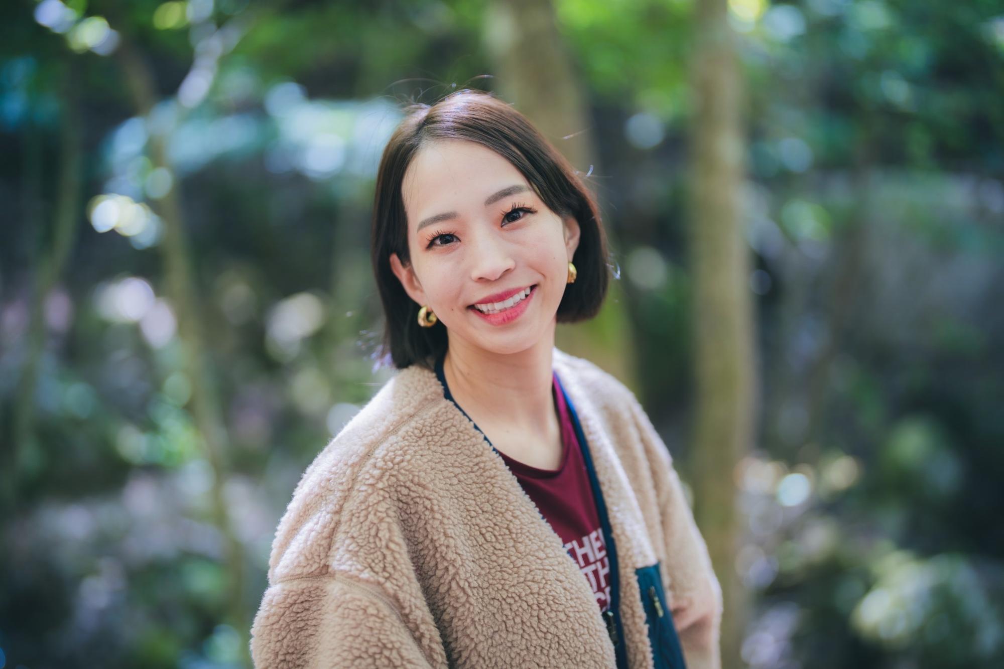 森や林のような緑の背景に、ボブヘアの野口啓代さんが白いボア素材の上着を着て、カメラに向かって微笑んでいる姿を明るい光の中で撮影した、人物にピントが合っている写真