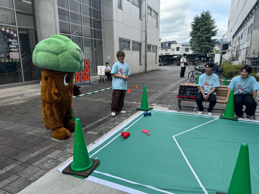 屋外の舗装された場所で、木の着ぐるみを着たキャラクターと3人の男性が、緑色のシートの上でボッチャのようなボールゲームをしているイベントの様子を写した写真