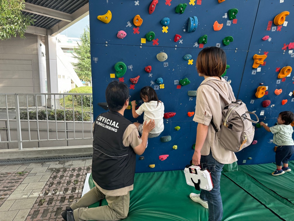 屋外に設置された青いクライミングウォールで、二人の幼児がカラフルなホールドに手をかけて登る練習をしており、その様子を一人の男性指導員と女性が見守っている写真