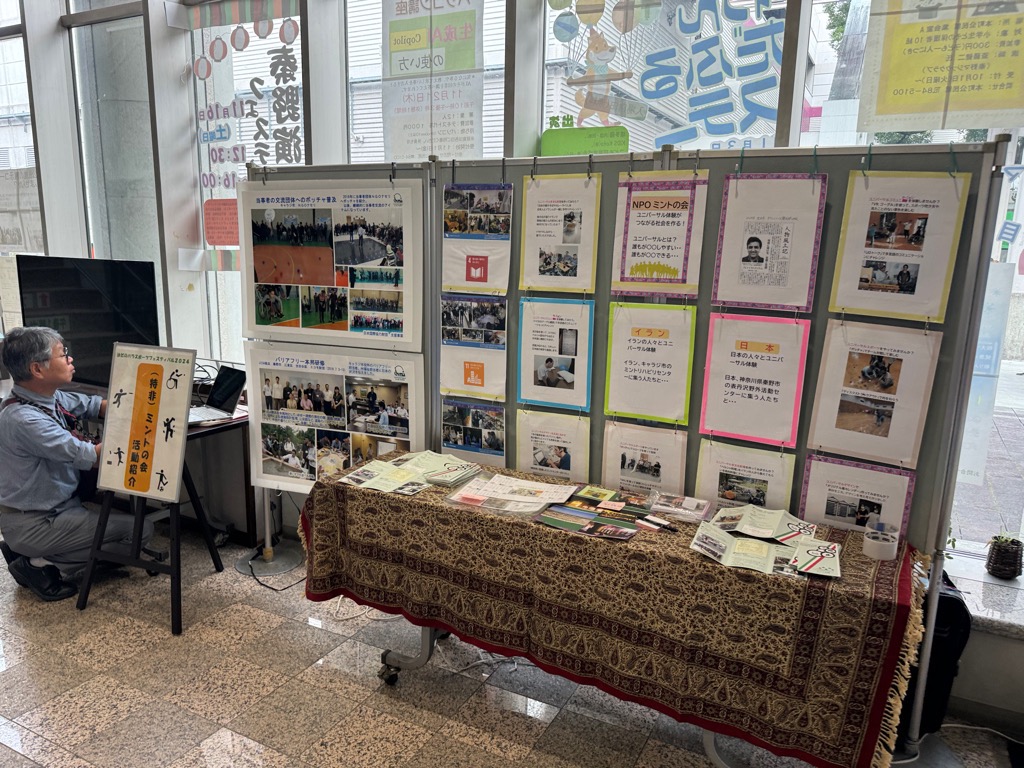 ガラス窓に面した屋内のブースで、男性が一人のぼりを立てたテーブルに着席し、壁面にNPOの活動を紹介する写真や文章のポスターが多数掲示されている様子の写真