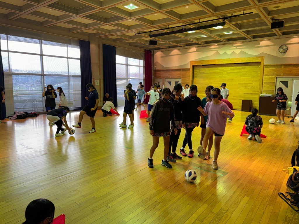 体育館のような広くて明るい室内で、木製の床の上で複数の学生がサッカーボールを使ったアクティビティに取り組んでおり、特に中央では目隠しをした女子学生がボールに触れようとしている写真