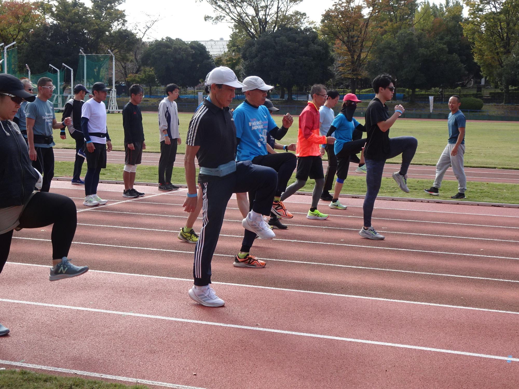 屋外の陸上競技場の赤いトラック上で、キャップをかぶり様々なスポーツウェアを着用した大人の集団が、右膝を高く上げ、手を振りながらトレーニングを行っている様子の写真