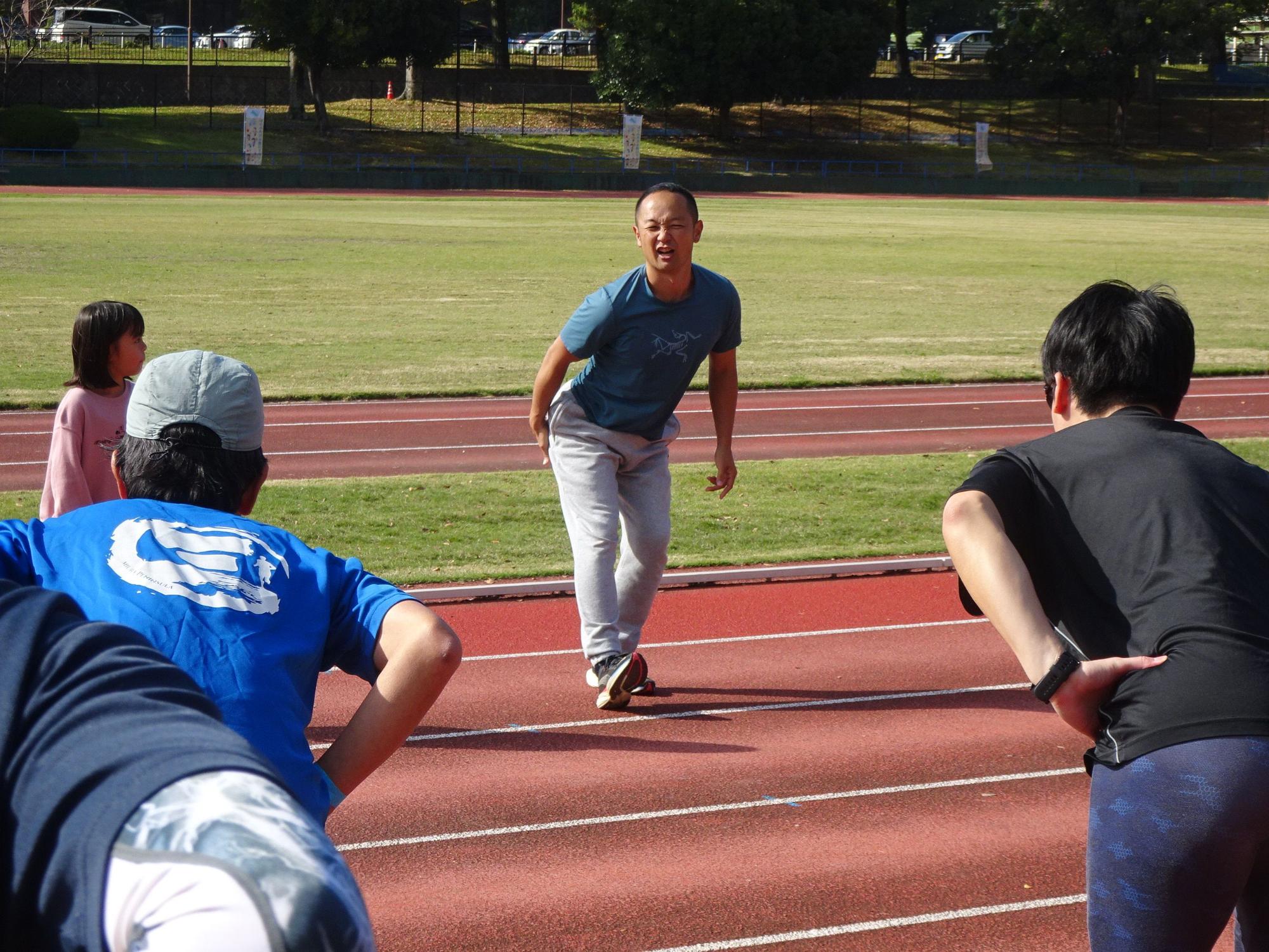 晴れた日の屋外の陸上競技場の赤いトラック上で、青いTシャツと灰色のスウェットパンツを着用した短髪の五味先生が、前かがみになって参加者たちに何かを教えており、周囲には熱心に耳を傾ける参加者たちの姿が見える指導風景の写真