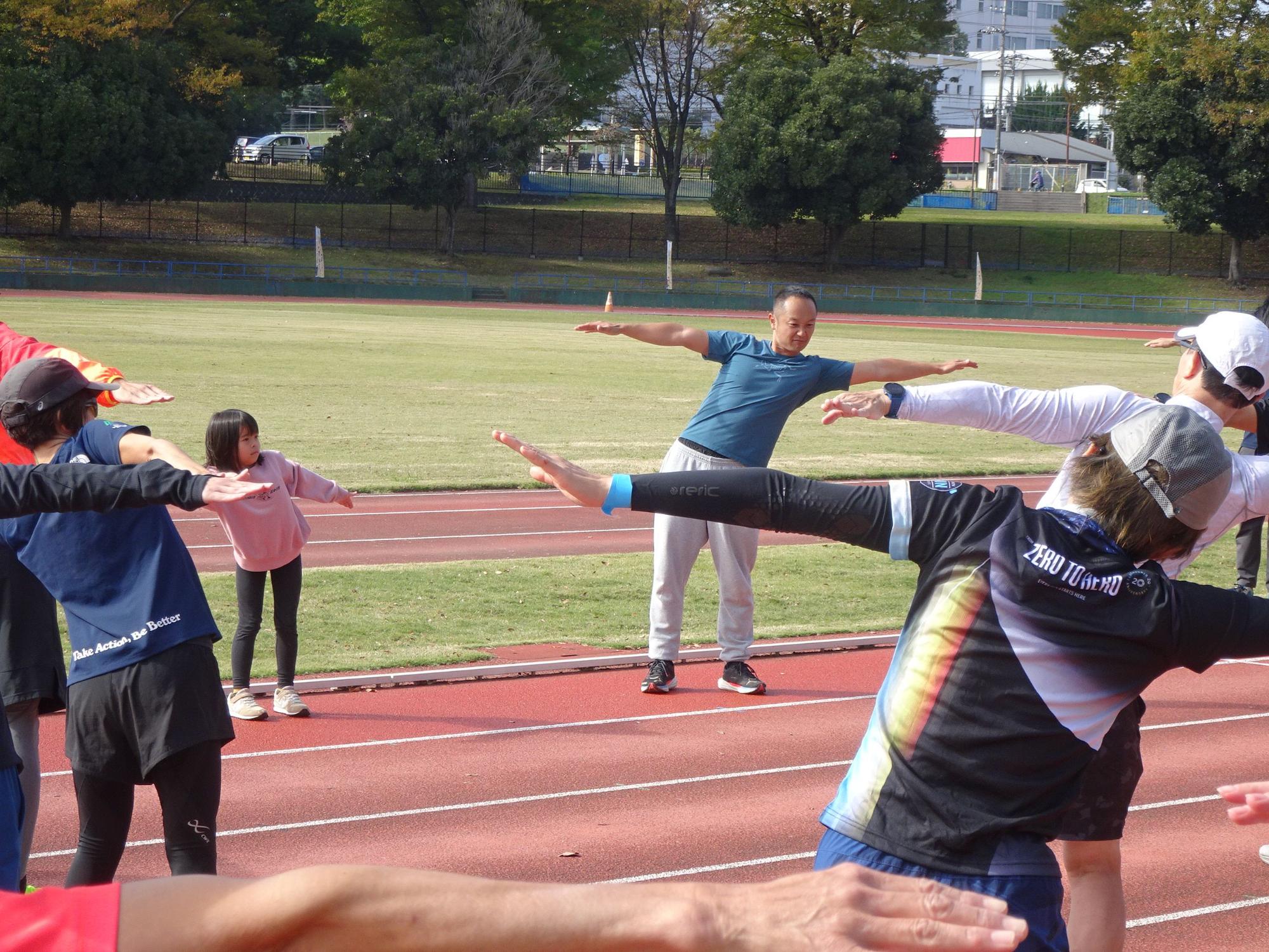 秋晴れの陸上競技場の赤いトラック上で、青いTシャツと灰色のスウェットパンツを着用した五味先生が、両手を広げて片足立ちのようなポーズをとり、数人の参加者も同様のストレッチをしている様子の写真