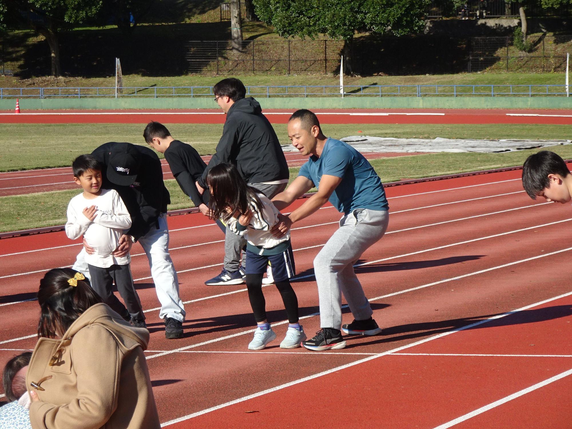 太陽が照りつける屋外の陸上競技場のトラックで、青いTシャツを着た男性が、黒い上着を着た大人や子供たちがいるグループの中で、少女の手を取りながら腰を落として何かを教えている様子の写真