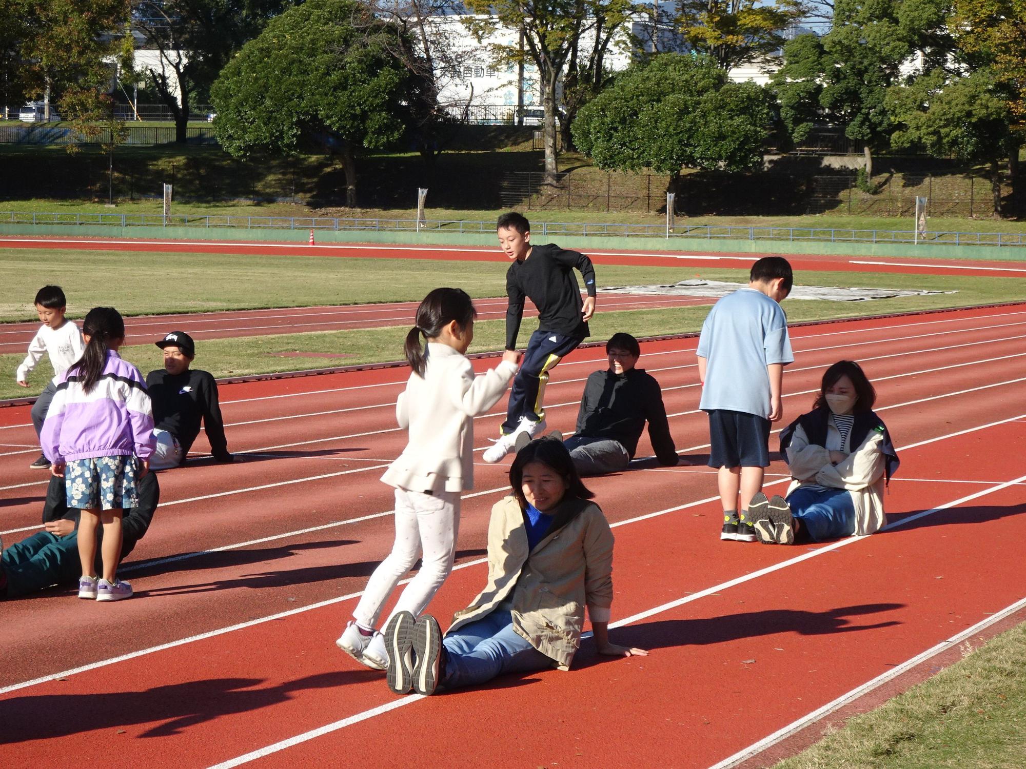 快晴の陸上競技場のトラックで、数人の大人と子供たちが赤い走路の上に座ったり、立ったりしている風景が写っており、特に白い上着を着た女の子が座っている女性の横で飛び跳ねている様子の写真