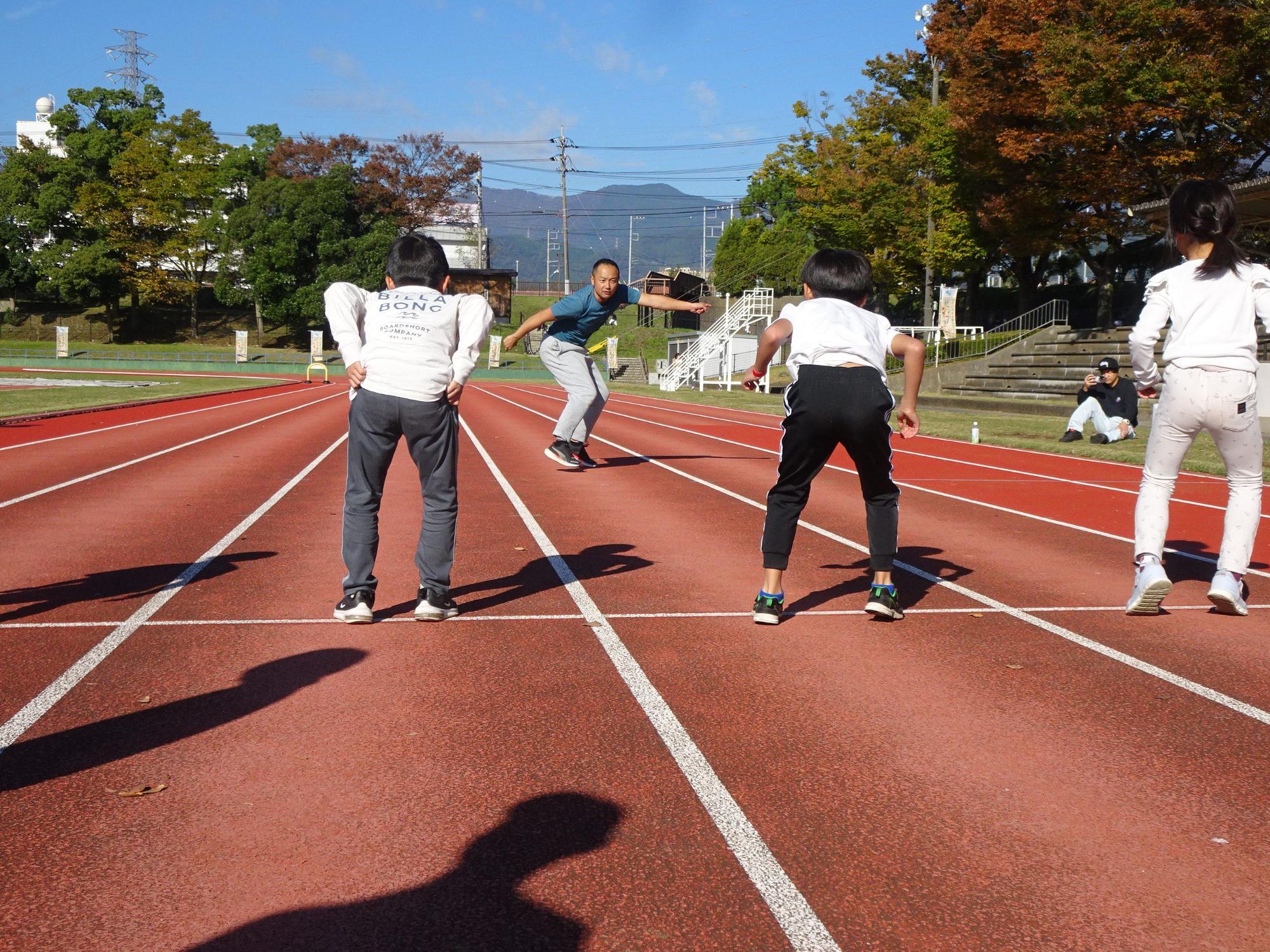 秋晴れの陸上競技場の赤いトラックで、青いTシャツと灰色のズボンを履いた男性が中央で両手を広げ、白い服を着た子供たちが彼に向かって走ろうとスタートラインに立っている写真