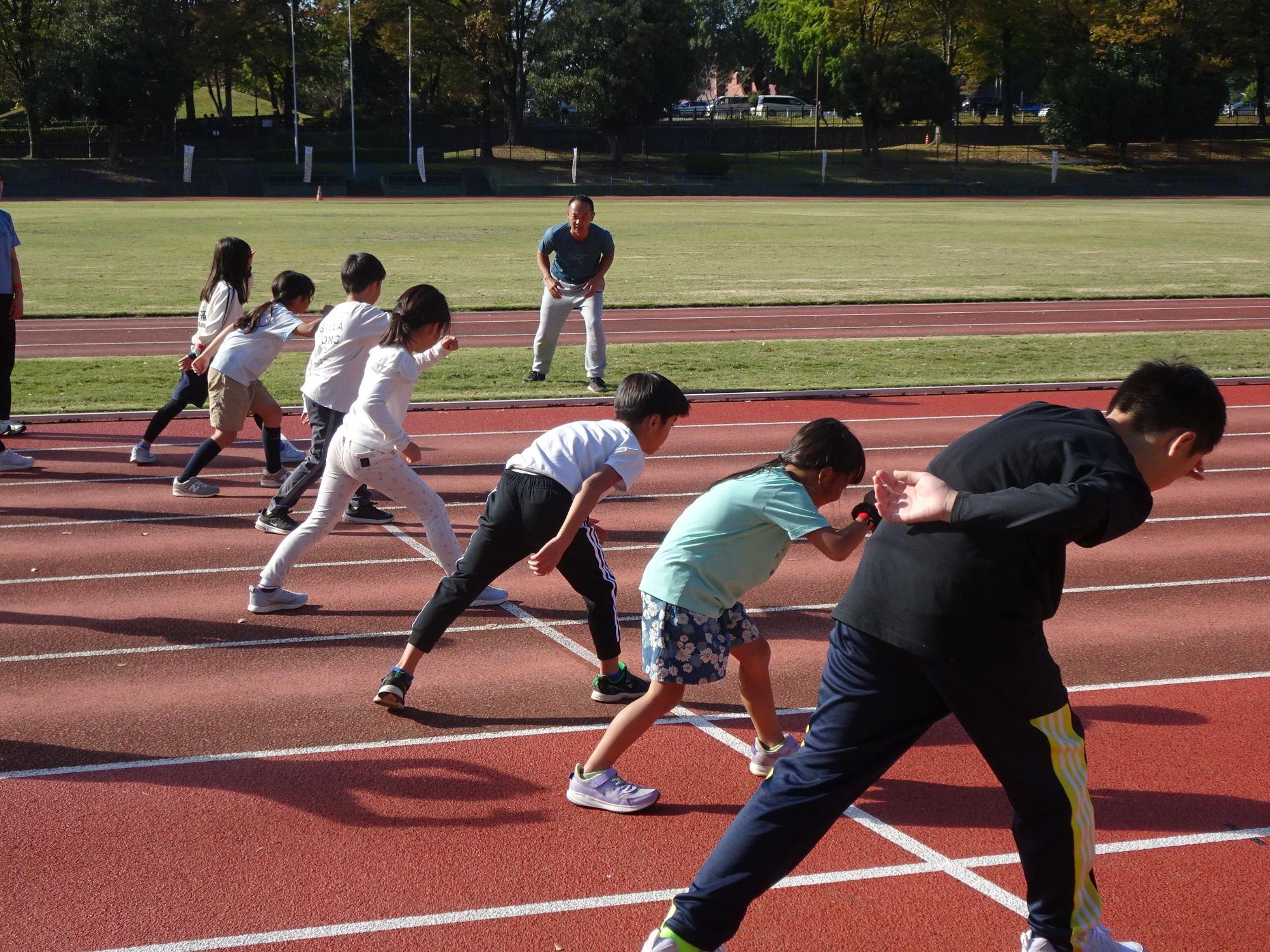 青いTシャツを着た男性が立って見守る中、白い線が引かれた陸上競技場の赤いトラックで、7人の子供たちが一列に並んでスタートの体勢をとり、前傾姿勢をとっている運動指導風景の写真
