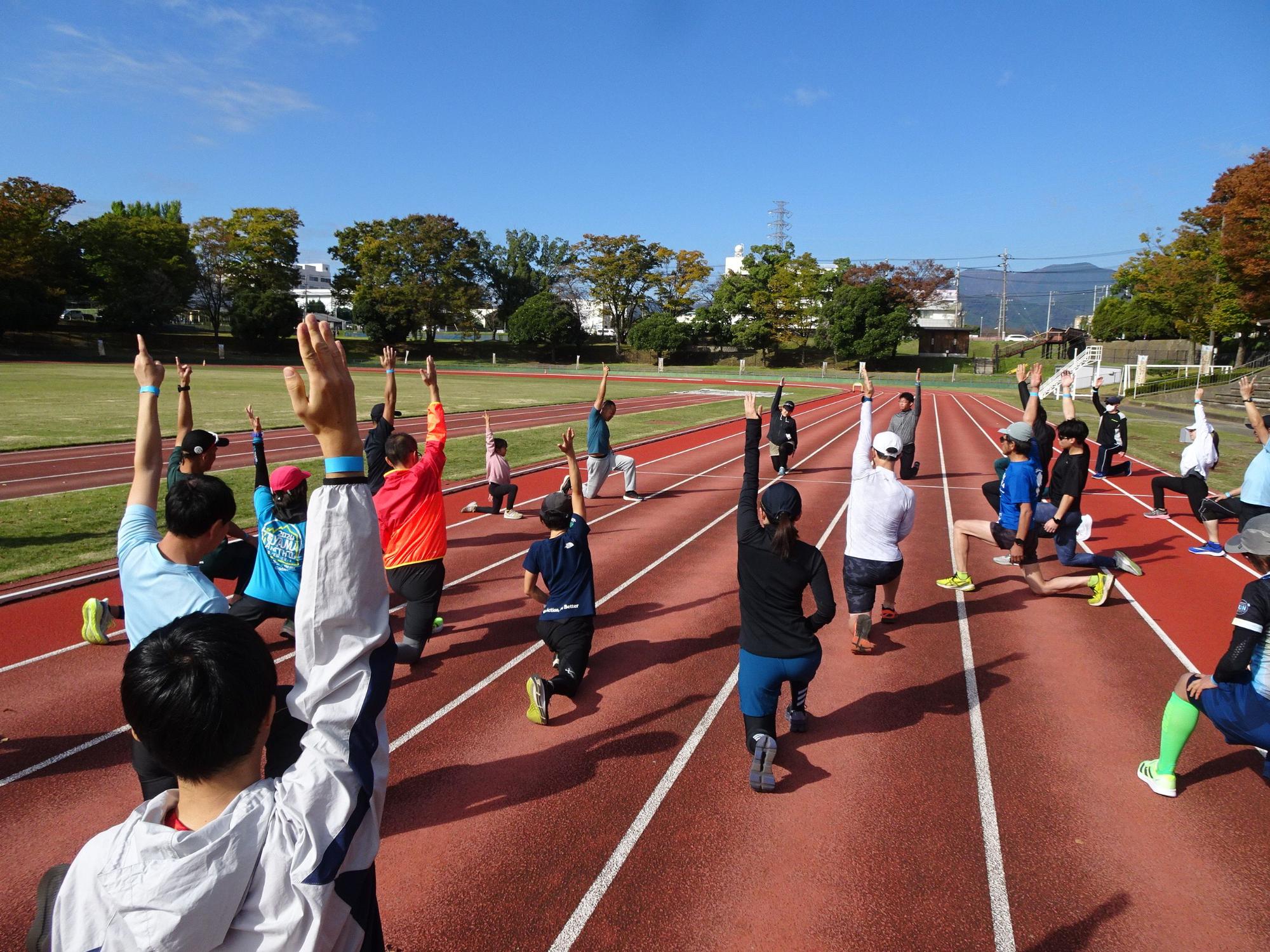 快晴の空の下、陸上競技場の赤いトラック上で、青いTシャツやカラフルなウェアを着た大人と子供の集団が、片膝をつきながら両手を高く空に伸ばして、準備運動のようなストレッチをしている様子の写真