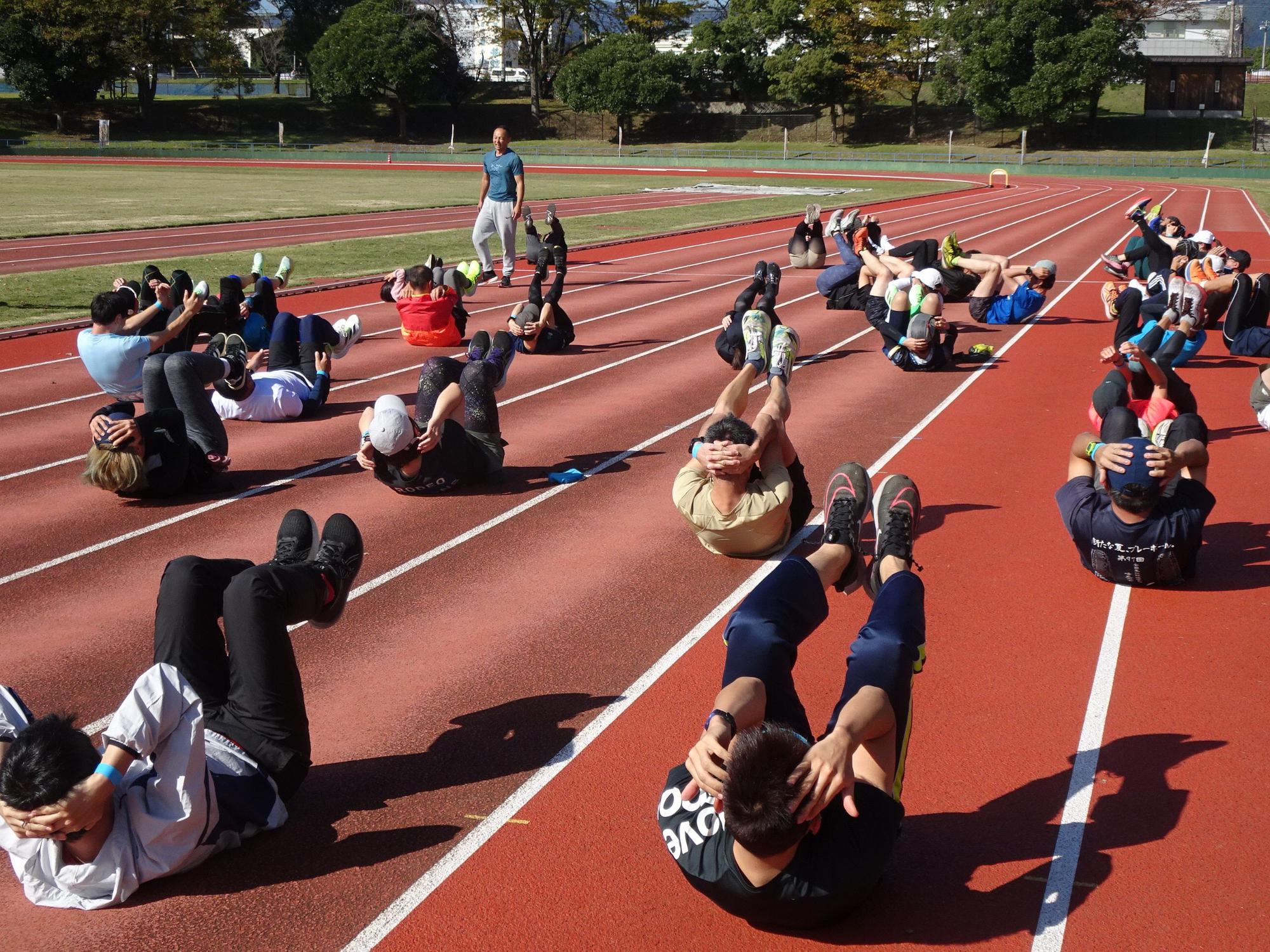 太陽が照りつける陸上競技場の赤いトラック上で、青いTシャツ姿の指導者らしき男性が見守る中、多くの参加者が仰向けになり、両膝を曲げて腹筋運動に取り組んでいるトレーニング風景の写真