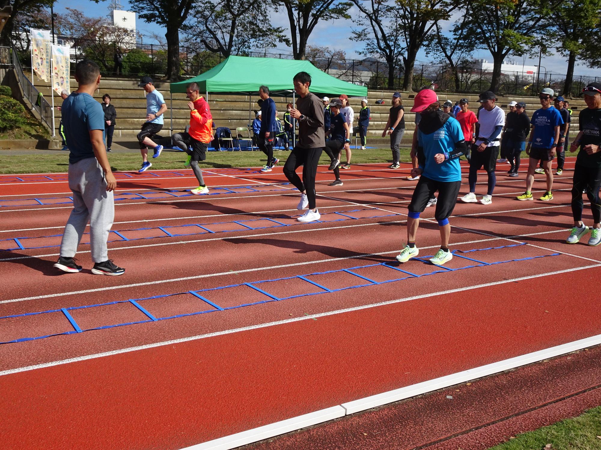 屋外の陸上競技場の赤いトラック上で、数人の参加者がアジリティラダーのような青い道具をまたぎながら腿上げやステップなどのトレーニングを行っており、奥には緑色のテントが見えるトレーニング風景の写真