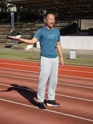 晴れた日の屋外の陸上競技場の赤いトラック上で、青いTシャツと灰色のスウェットパンツを着用した短髪の五味 宏生さんが、手に黒いランニングシューズを乗せて高く掲げ、口を開けて何かを説明している様子の写真