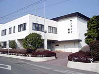 白を基調とした二階建ての建物の正面玄関と、手入れされた植え込みが並ぶ様子を捉えたサンライフ鶴巻の外観写真