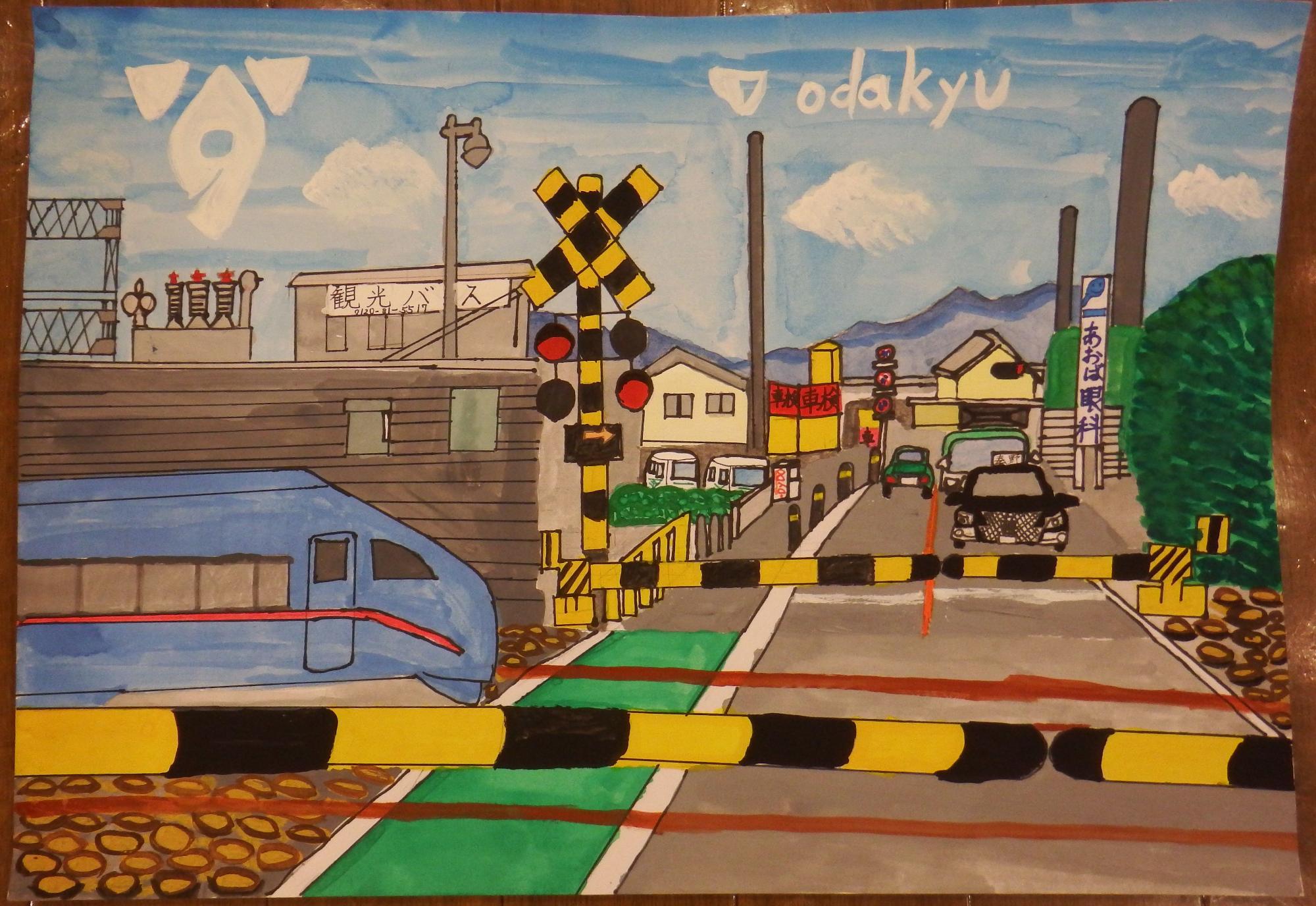 kyouikutyou「odakyu」のロゴが浮かぶ空と山を背景に、遮断機がおりた道路に順番に車が停車し、警報機が鳴っている線路を青い電車が通過する様子が描かれた踏切の風景を、道路標識や眼科の看板に建物、車が遠近法を使って描かれている秦野市教育長賞受賞作品の写真