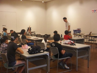 たくさんの子供たちが机に座って部屋の前方に立つ男性の話を聞いており、一人の女の子が手を挙げて質問している、創作講座の様子の写真