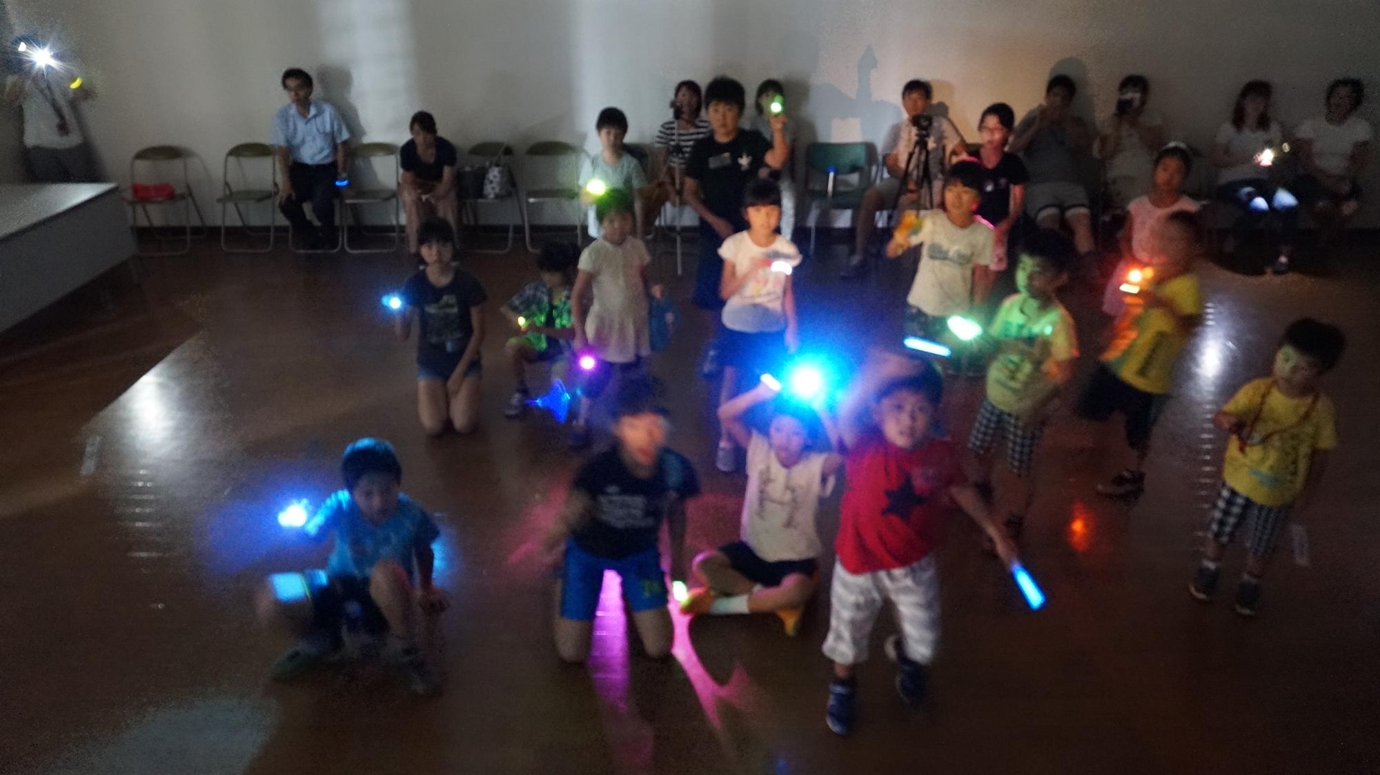 薄暗い部屋の中で、たくさんの子どもたちが手に持った光る棒を振り回して楽しんでおり、後ろには数人の大人が座っている様子を捉えた写真