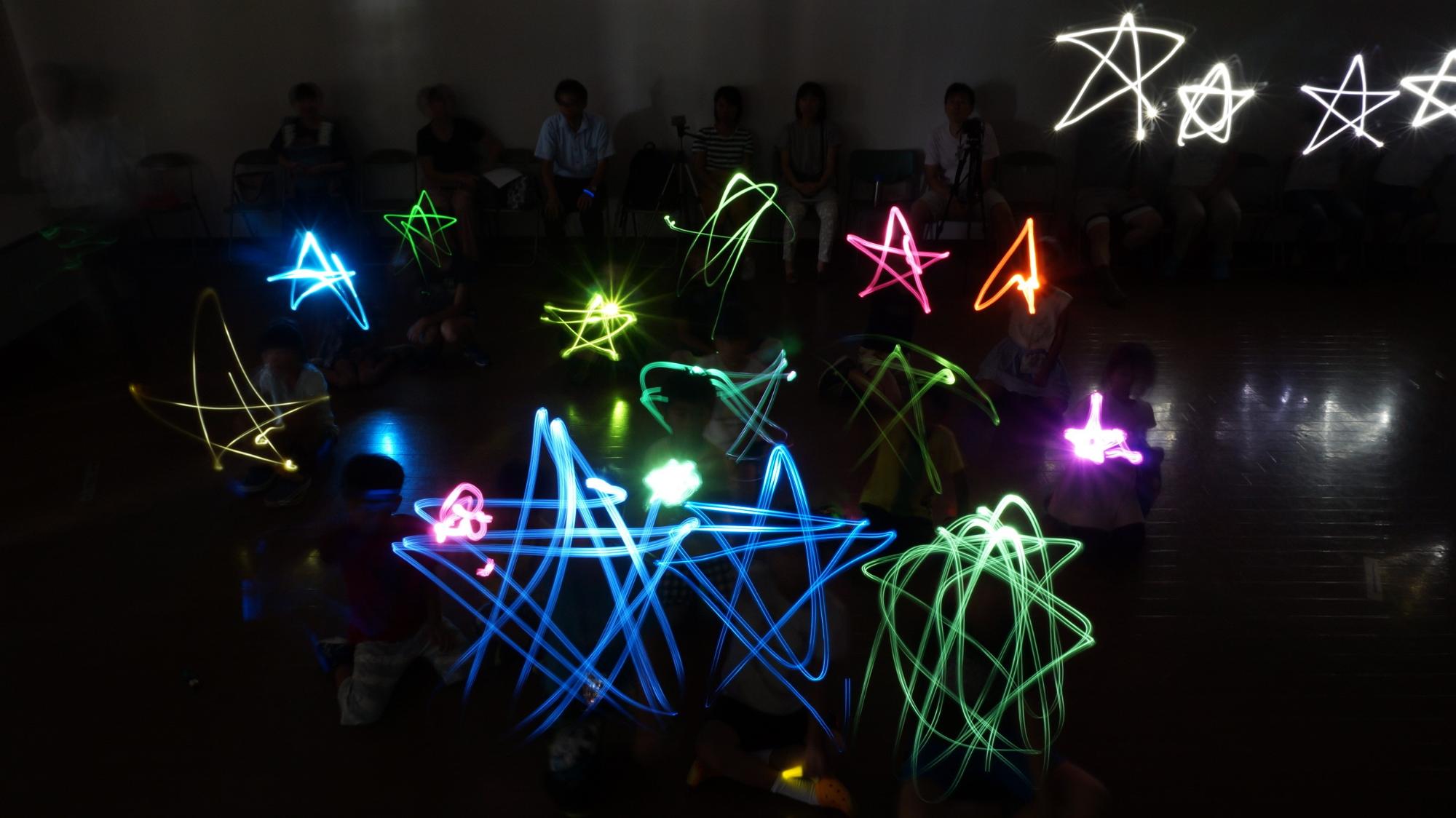 暗い部屋の中で、たくさんの子どもたちが手に持った光る棒で星の形を作って楽しんでおり、後ろには数人の大人が座っている様子が薄っすら写っている様子を捉えた写真