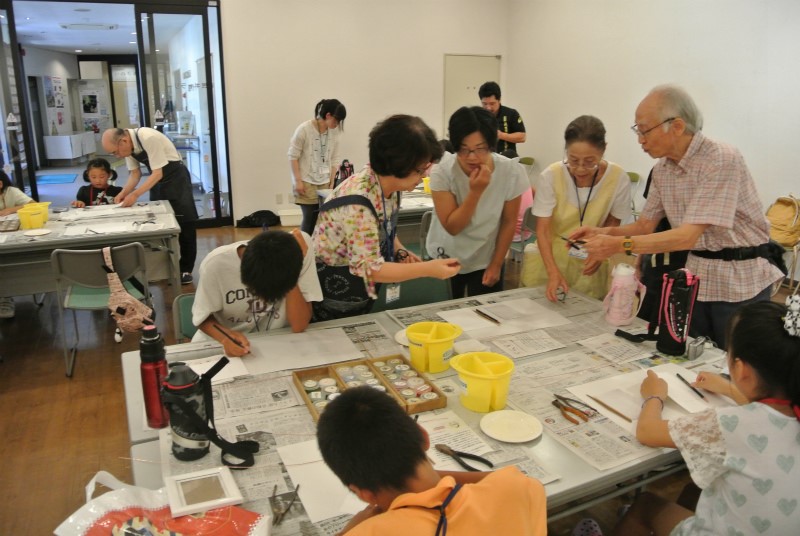 子供たちが絵を描いている新聞紙を広げた机の周りで、3人の講師が水色のシャツを着た母親に説明をしている様子の写真