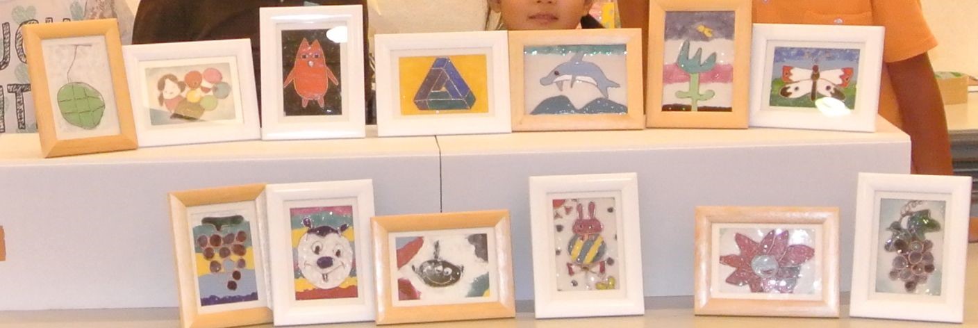 イルカや蝶、葡萄やメロンなどの果物に花など、子供たちが制作したカラフルな作品が、木製や白いフレームに入れられて、机の上に二段に並べられている写真