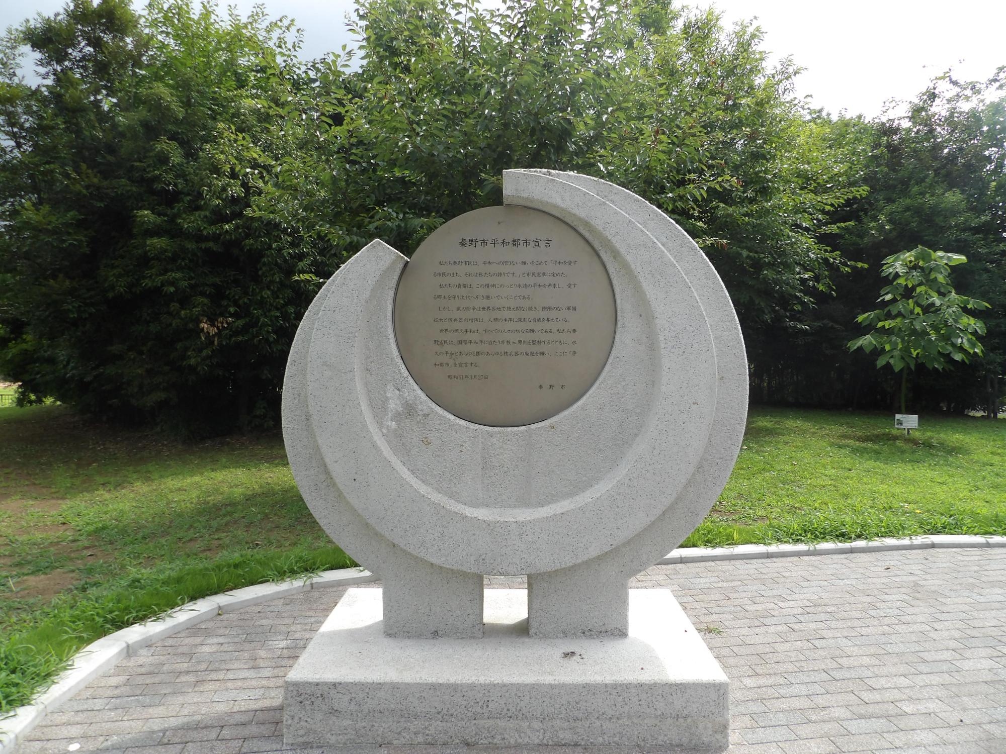 緑豊かな木々や草に囲まれた広場に、四角形の土台の上に円形の御影石のような質感の石碑が設置されている平和都市宣言記念碑の写真