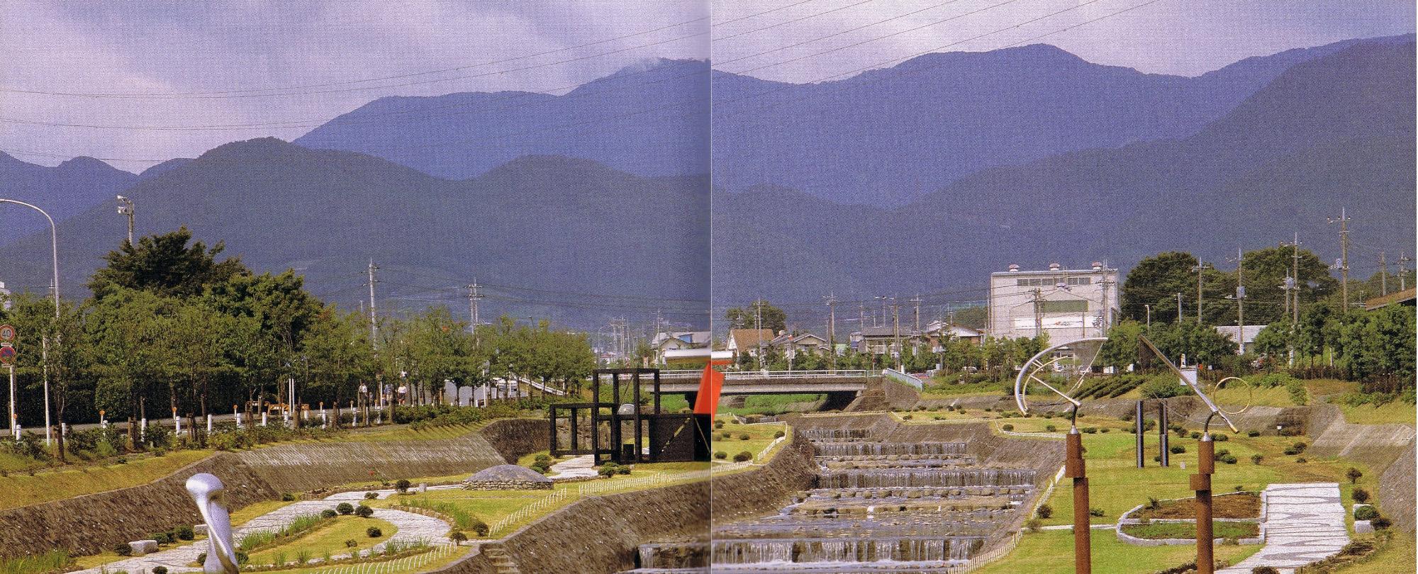 青い山脈を背景に、コンクリートで整備された川岸に沿って階段状の小滝と緑地、散策路が広がり、現代的なオブジェが点在する公園の全景を捉えた風景写真