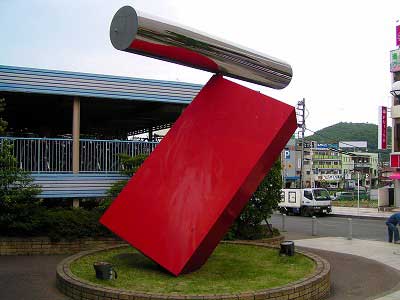 広場にある芝生がの植え込みに設置された、赤く傾いた四角柱の土台の上に、今にも倒れそうな斜めに載せられた銀色に輝く円筒形の物体で構成される抽象的なオブジェの写真