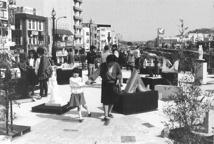 背景には商店やビルが立ち並ぶ都市の景観が広がる歩行者広場のような場所で、大人や子供を含む複数の人々が様々な抽象的な現代アートの彫刻を鑑賞している様子を捉えた白黒写真