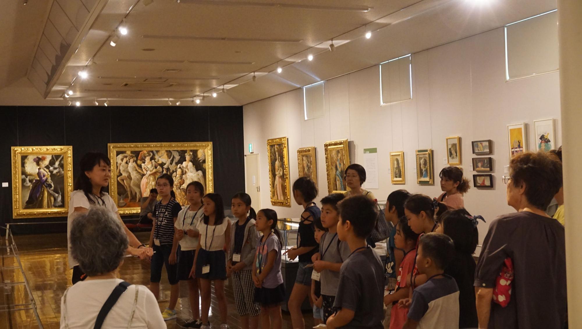 たくさんの親子連れの参加者が大小様々な絵画作品が展示されている美術館の展示室に集まり、白い服を着た女性スタッフの説明を子供たちが熱心に聞いている様子の写真