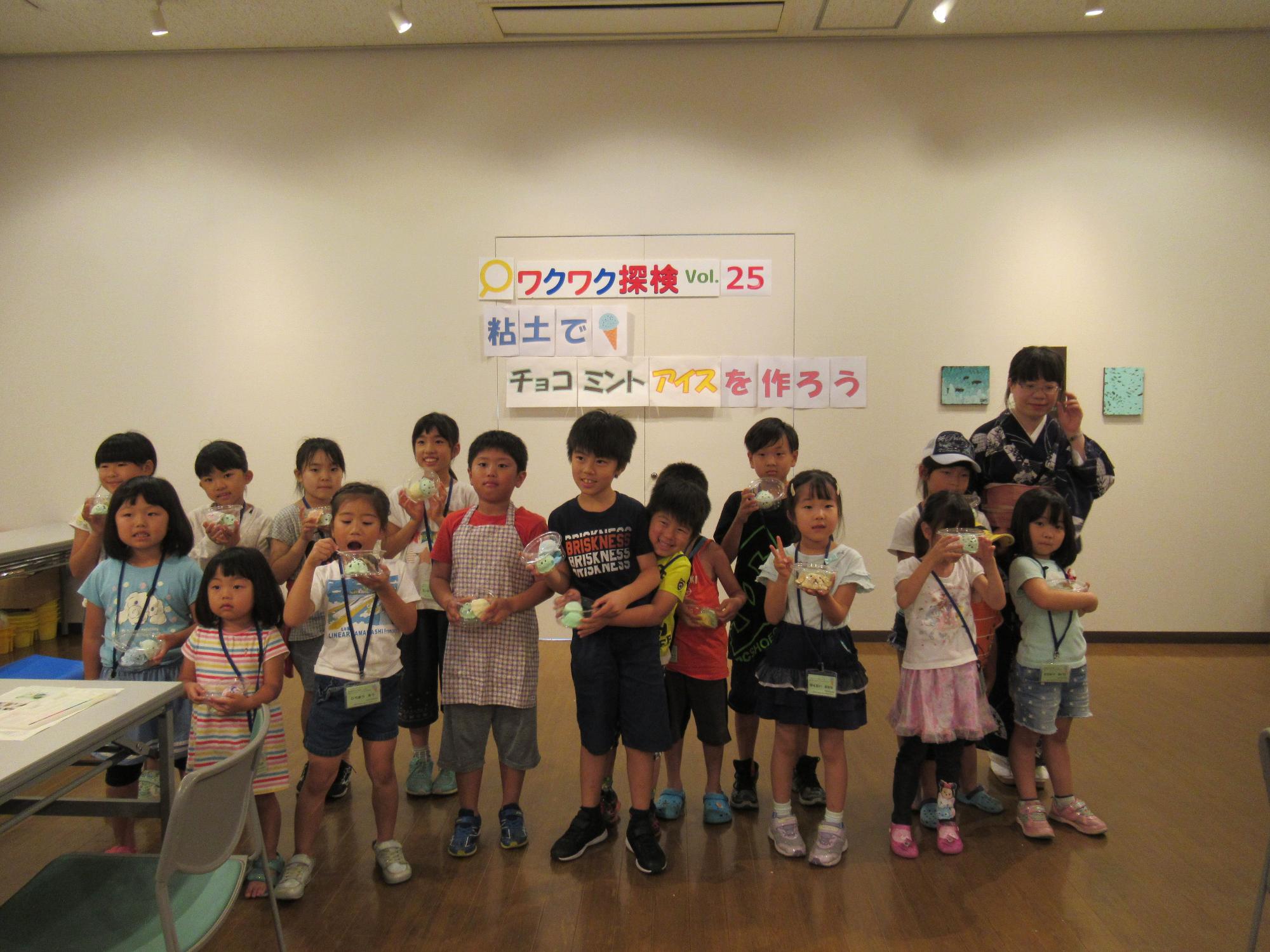「ワクワク探検 Vol.25 粘土でチョコミンアイスを作ろう」と書かれた白い壁の前で、着物を着た女性や、参加した子供たちが自作のアイス作品を手に持ち、マスクを着用せずに記念撮影をしている集合写真